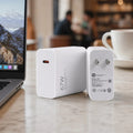 Two white 67W USB-C power adapters on a light marble tabletop in a cafe setting, with a laptop on the left and a white coffee cup on the right, blurred in the background.